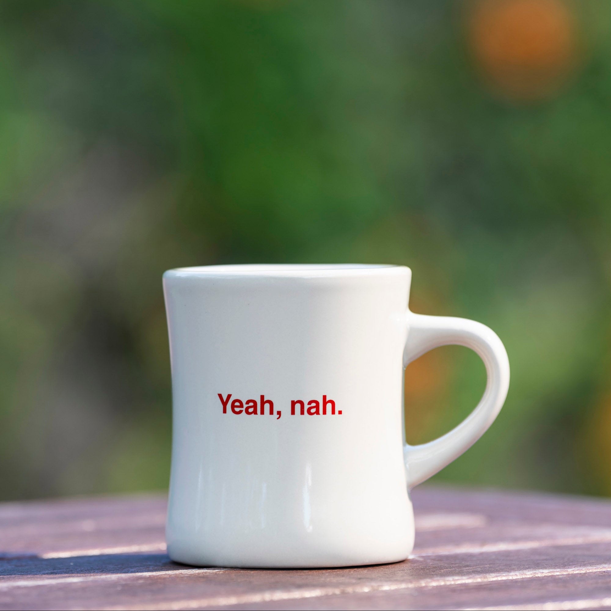 White mug with 'Yeah, nah.' text on a blurred natural background