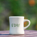 White mug with 'G'DAY' text on a wooden surface with a blurred green background