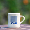 White mug with 'NOOSA' branding on a wooden surface with a blurred natural background