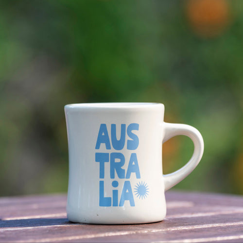White mug with 'Australia' printed in blue on a wooden surface with a blurred natural background